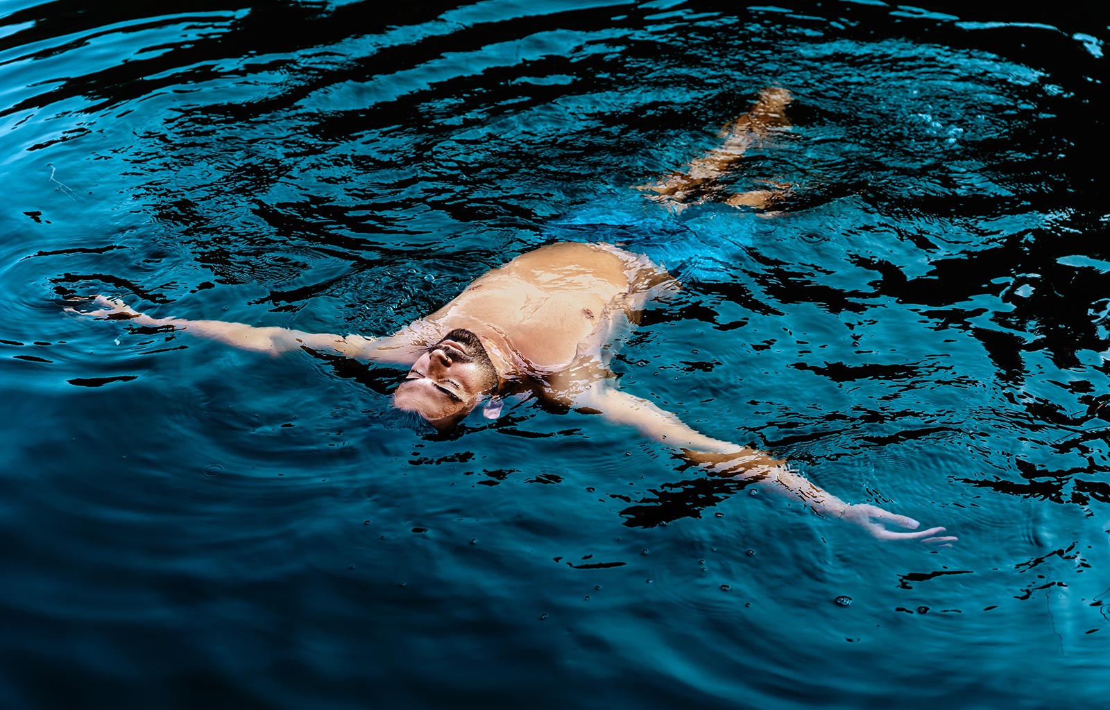 A young bearded man floats calmly on the surface of a dark blue body of water, with his arms outstretched and his eyes closed. 