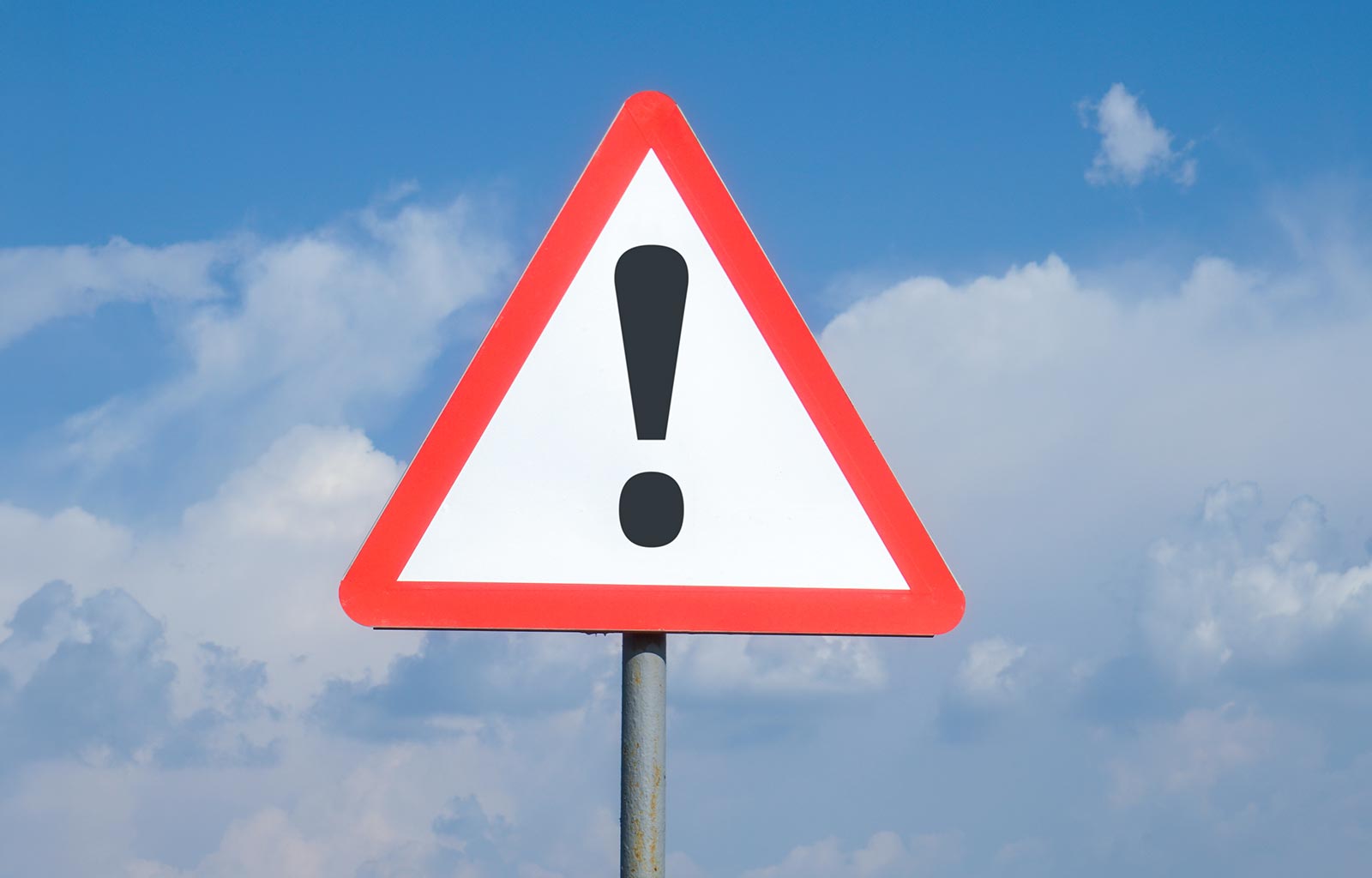  An exclamation mark on a triangular road sign. The background is a blue sky with clouds. The sign indicates a warning, danger, attention required.